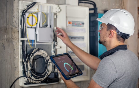 Man, an electrical technician working in a switchboard with fuses, uses a tablet.